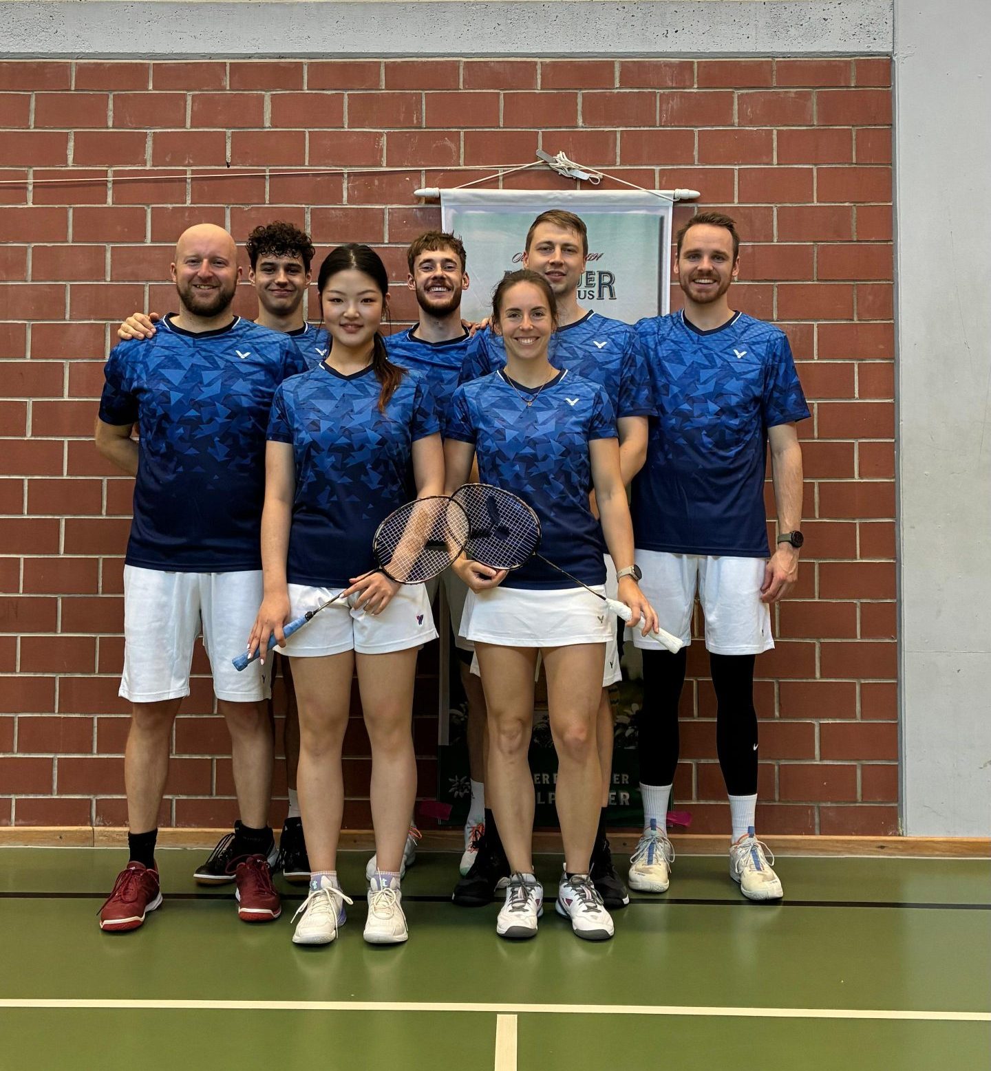 1. Mannschaft Eine Badmintonmannschaft mit sieben Personen steht in einer Sporthalle vor einer Backsteinwand. Alle tragen blaue Trikots und weiße Shorts. Zwei Spielerinnen in der vorderen Reihe halten Badmintonschläger, während die fünf Männer hinter ihnen stehen und lächeln. Der Boden ist ein grünes Badmintonfeld, und hinter der Gruppe hängt ein Banner an der Wand.