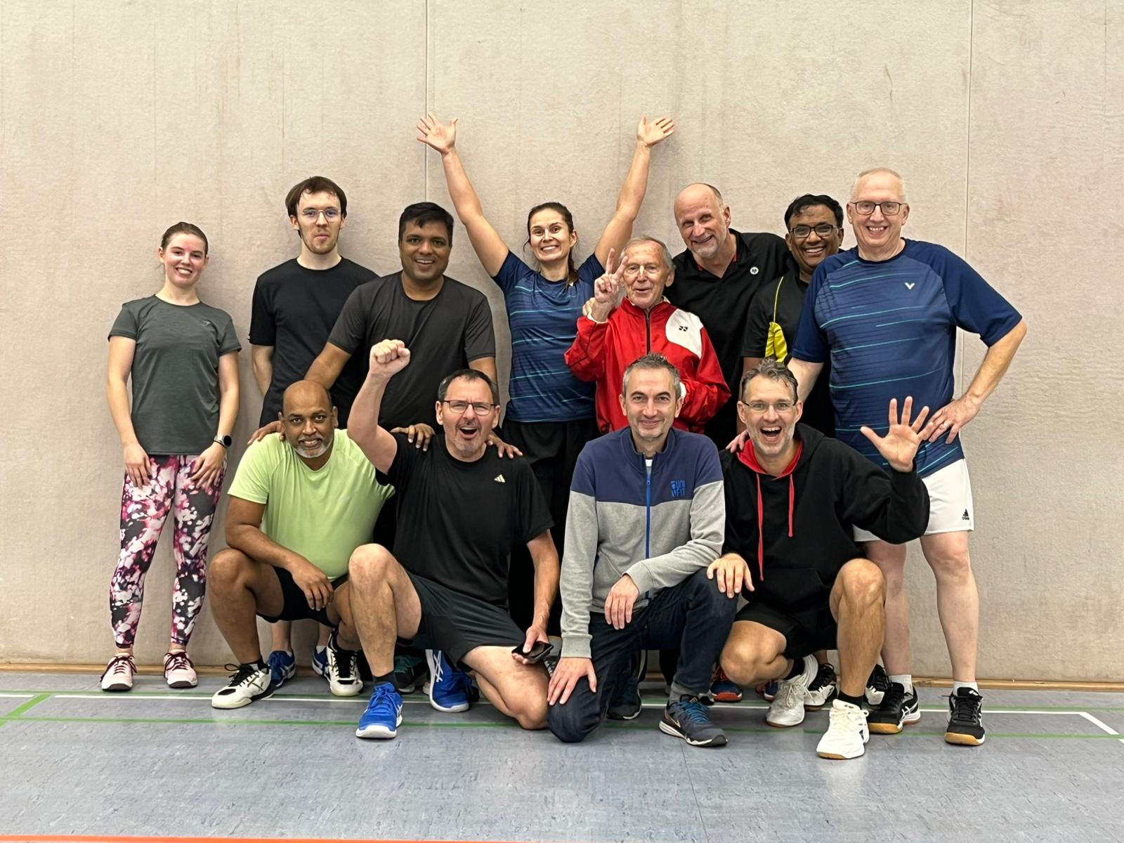 Gruppenfoto einer fröhlichen Badminton-Trainingsgruppe in einer Sporthalle. Dreizehn Personen unterschiedlichen Alters stehen und knien in zwei Reihen. Einige lachen, andere winken oder zeigen jubelnde Gesten. Alle tragen Sportkleidung und stehen vor einer hellen Hallenwand.