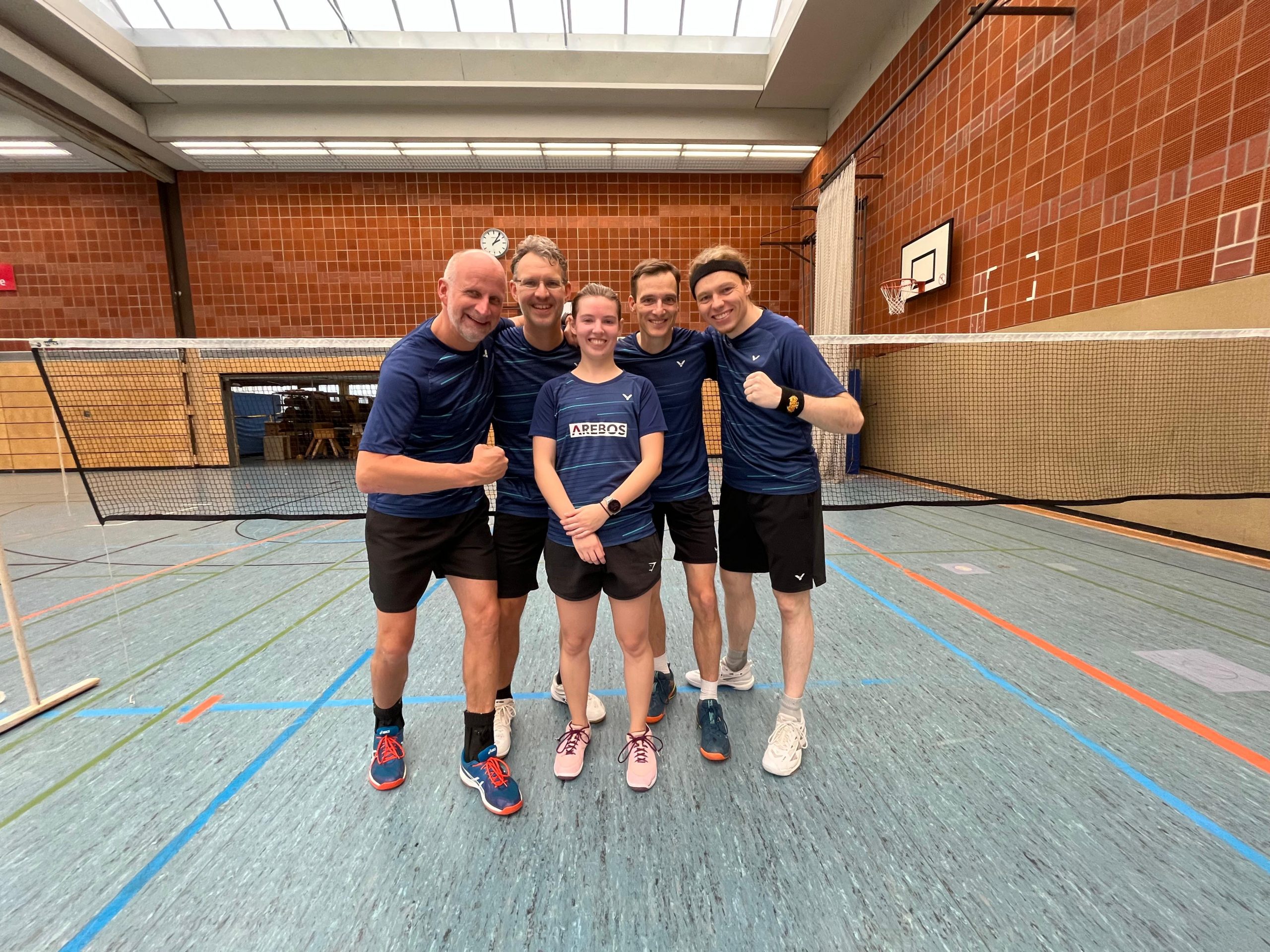 Fünf Badmintonspielerinnen und -spieler stehen in einer Sporthalle vor einem Badmintonnetz. Alle tragen blaue Trikots und schwarze Shorts, lächeln in die Kamera und wirken fröhlich und motiviert.