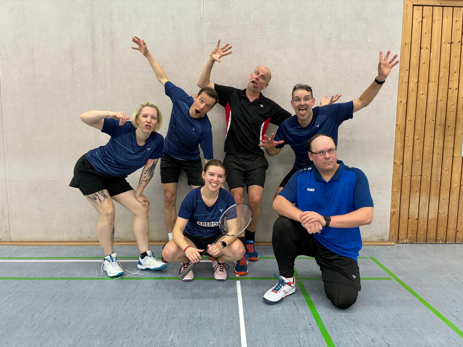 Sechs Badmintonspielende in einer Sporthalle posieren vor einer Hallenwand; fünf stehen mit lustigen Gesten und erhobenen Armen, eine Person kniet vorn mit Badmintonschläger.