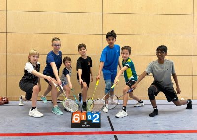 Acht Kinder stehen nebeneinander in einer Sporthalle und halten Badmintonschläger nach vorne. Sie tragen Sportkleidung und nehmen eine Ausfallschritt-Position ein. Vor ihnen steht eine Punktezähltafel auf dem Hallenboden, im Hintergrund ist eine helle Holzpaneelwand zu sehen.“